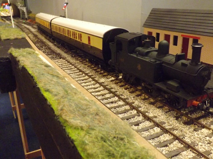A detailed model train set with a steam locomotive pulling several passenger cars along a track. The setup includes realistic tracks, grass-covered embankments, and signaling equipment. A small building with a red roof is visible alongside the tracks.