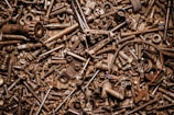 A dense collection of various metal bolts, screws, nuts, and mechanical parts in a chaotic arrangement. The image conveys a sense of clutter and industrial material, with a variety of metallic textures and shades.
