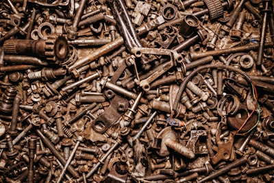 A dense collection of various metal bolts, screws, nuts, and mechanical parts in a chaotic arrangement. The image conveys a sense of clutter and industrial material, with a variety of metallic textures and shades.