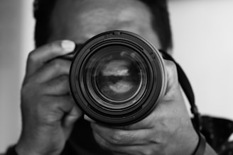 A close-up black and white photograph of a person holding a camera, with the focus on the camera lens. The hands are visible, gripping the camera securely, and the complexion of the person is obscured due to depth of field.