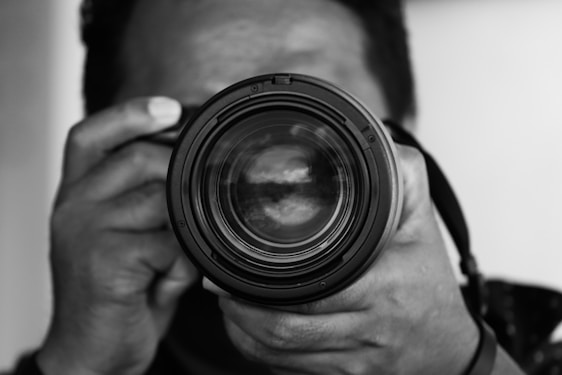 A close-up black and white photograph of a person holding a camera, with the focus on the camera lens. The hands are visible, gripping the camera securely, and the complexion of the person is obscured due to depth of field.