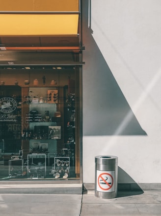 A storefront with a large window display showcasing various items. To the right, a metal trash can stands against a white wall, featuring a no smoking sign. A yellow awning casts a shadow on the wall and pavement, adding a geometric play of light and shade.