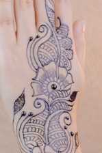 A hand adorned with intricate henna designs featuring floral and paisley motifs. The design is detailed with curved lines and dot accents, creating an elegant and artistic look.