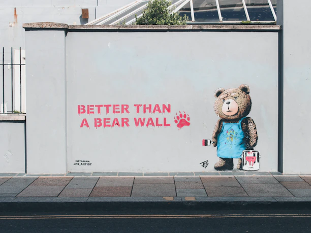 A wall mural features a bear wearing a blue apron holding a paintbrush and a bucket of paint. The text next to the bear reads 'Better than a bear wall' in red paint with a bear paw print. The scene is set on a light grey wall along a sidewalk.