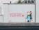 A wall mural features a bear wearing a blue apron holding a paintbrush and a bucket of paint. The text next to the bear reads 'Better than a bear wall' in red paint with a bear paw print. The scene is set on a light grey wall along a sidewalk.