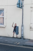A street corner featuring wall art depicting a Roman soldier holding a sword and shield. The artwork is painted on a light-colored wall adjacent to a window with a visible curtain. A metal pole with street signs is positioned next to the painting, and the ground is covered with gray pavement.