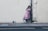 A mural of a young girl wearing a pink dress and white sneakers, painted on an urban wall, seemingly hiding or playing against a drainpipe. A paint can is tipped over on the pavement next to her.