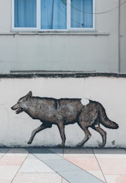 A mural of a wolf is painted on a pale wall below a window with blue curtains. The lifelike artwork is positioned just above a tiled pavement, creating a realistic effect as if the wolf is walking across the tiles.