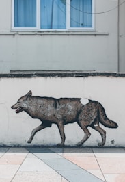 A mural of a wolf is painted on a pale wall below a window with blue curtains. The lifelike artwork is positioned just above a tiled pavement, creating a realistic effect as if the wolf is walking across the tiles.