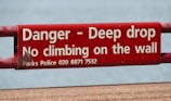 A red warning sign with white text stating 'Danger - Deep drop, No climbing on the wall.' It includes a contact number for Parks Police.
