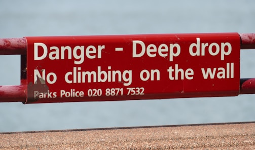 A red warning sign with white text stating 'Danger - Deep drop, No climbing on the wall.' It includes a contact number for Parks Police.