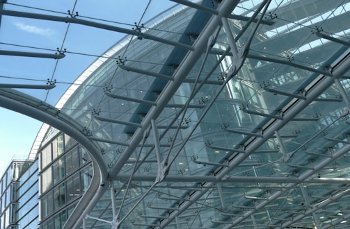 A complex structure of glass and steel forms an architectural feature, with overlapping panes of clear glass supported by a grid of metal beams. The design is both geometric and modern, showcasing intricate engineering and design. The sky is visible through the transparent sections, adding to the airy and open feel of the construction.