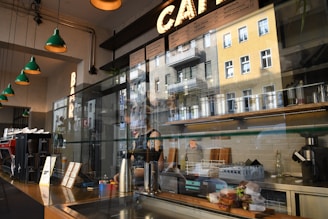 The image captures the interior of a modern cafe with a view reflecting the outside urban architecture through the glass. The cafe counter features various coffee-making equipment, menu boards, and small plants, creating a warm ambiance with green pendant lights hanging from the ceiling.