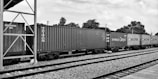 A set of freight train cars carrying shipping containers is stationed on railway tracks. The scene includes several branded containers, featuring names like Triton, Hamburg Sud, and Maersk. The background consists of trees and utility poles under a partially cloudy sky. The setting appears to be an open railway yard or station.