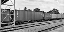 A set of freight train cars carrying shipping containers is stationed on railway tracks. The scene includes several branded containers, featuring names like Triton, Hamburg Sud, and Maersk. The background consists of trees and utility poles under a partially cloudy sky. The setting appears to be an open railway yard or station.