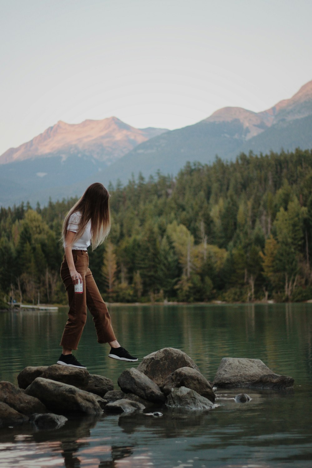 Mujer que pisa las rocas grises cerca de la montaña que mira al lago durante el día