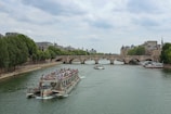 A river scene featuring several boats navigating through the water. In the foreground, a large tour boat is filled with passengers. Behind it, a stone arch bridge spans the river, connecting two banks lined with trees and historic buildings. The sky is partially cloudy, casting a soft light over the scene.