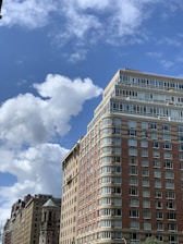 Tall residential buildings with large glass windows are set against a bright blue sky with scattered white clouds. The architecture features a mix of brick and stone, with balconies visible on the top floors.