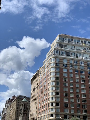 Tall residential buildings with large glass windows are set against a bright blue sky with scattered white clouds. The architecture features a mix of brick and stone, with balconies visible on the top floors.