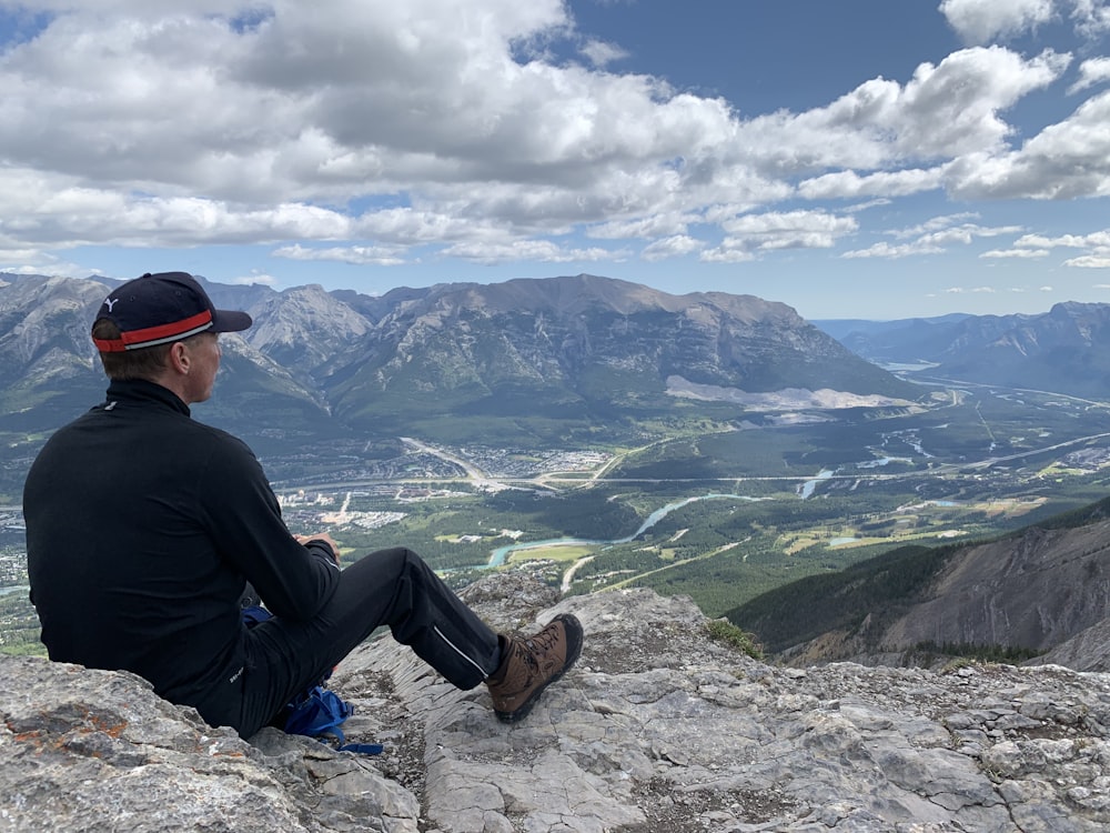 man sitting on rock cliff under white clouds