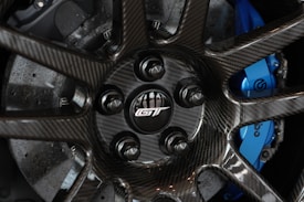 A close-up view of a carbon fiber wheel featuring a prominently displayed lug nut section with a GT logo in the center. The blue brake caliper, marked with a recognizable automotive brand logo, is partially visible through the wheel spokes.