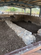 A covered archaeological excavation site featuring ancient stone walls arranged in a rectangular formation. The site is sheltered by a modern structure with a wooden and metal roof. Surrounding the site is a wooden fence, and in the background, there are green trees and distant hills under a cloudy sky.