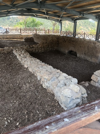 A covered archaeological excavation site featuring ancient stone walls arranged in a rectangular formation. The site is sheltered by a modern structure with a wooden and metal roof. Surrounding the site is a wooden fence, and in the background, there are green trees and distant hills under a cloudy sky.