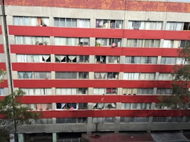 A multi-story residential building with a red and gray facade. Numerous windows are visible, some covered with curtains and others displaying various personal items like plants. Satellite dishes are mounted on some of the balconies. The building's exterior appears worn and aged, with visible signs of weathering.