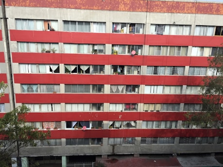 A multi-story residential building with a red and gray facade. Numerous windows are visible, some covered with curtains and others displaying various personal items like plants. Satellite dishes are mounted on some of the balconies. The building's exterior appears worn and aged, with visible signs of weathering.