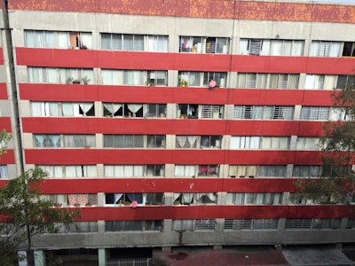 A multi-story residential building with a red and gray facade. Numerous windows are visible, some covered with curtains and others displaying various personal items like plants. Satellite dishes are mounted on some of the balconies. The building's exterior appears worn and aged, with visible signs of weathering.