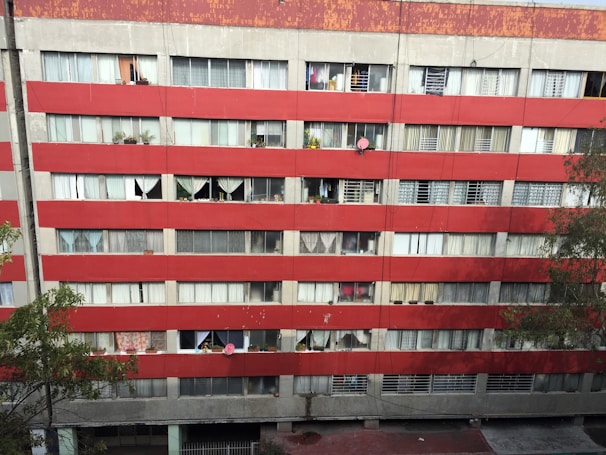 A multi-story residential building with a red and gray facade. Numerous windows are visible, some covered with curtains and others displaying various personal items like plants. Satellite dishes are mounted on some of the balconies. The building's exterior appears worn and aged, with visible signs of weathering.