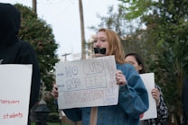 A group of protestors hold signs advocating for free education and opposing cuts and fees. One prominent protestor has taped their mouth shut and holds a sign that reads 'NO CUTS NO FEES EDUCATION MUST BE FREE.' The protest takes place outdoors, with trees and a building visible in the background.