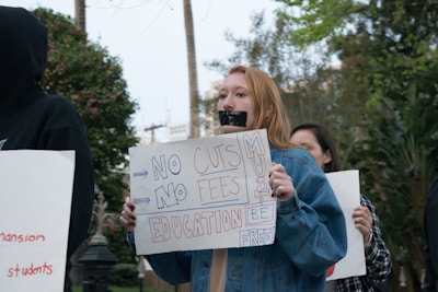 A group of protestors hold signs advocating for free education and opposing cuts and fees. One prominent protestor has taped their mouth shut and holds a sign that reads 'NO CUTS NO FEES EDUCATION MUST BE FREE.' The protest takes place outdoors, with trees and a building visible in the background.