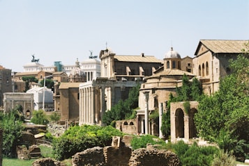 A historical cityscape featuring ancient ruins with stone columns and arches surrounded by lush greenery. The architecture includes domed buildings and long colonnades, creating an impression of a classical-era setting. In the background, there's a modern monument with statues and more contemporary structures.
