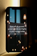 A smartphone leaning against a wall displays a message about conserving energy by turning off unused power points and lights. The screen background features hanging light bulbs, some lit and others dim, casting a warm glow against a dark and possibly urban skyline.