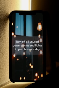 A smartphone leaning against a wall displays a message about conserving energy by turning off unused power points and lights. The screen background features hanging light bulbs, some lit and others dim, casting a warm glow against a dark and possibly urban skyline.