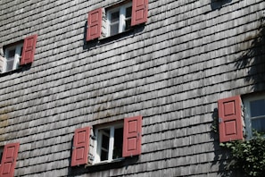A building facade with traditional wooden shingle siding and multiple windows, each with red shutters. The windows are open, allowing a view of the green foliage inside. There are five windows with the sunlight casting shadows, adding depth to the textured surface.