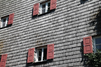 A building facade with traditional wooden shingle siding and multiple windows, each with red shutters. The windows are open, allowing a view of the green foliage inside. There are five windows with the sunlight casting shadows, adding depth to the textured surface.