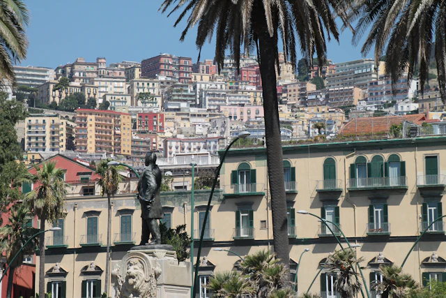 A statue and palm trees in the foreground with the dense, colourful hillside apartment blocks of Vomero rising behind.
