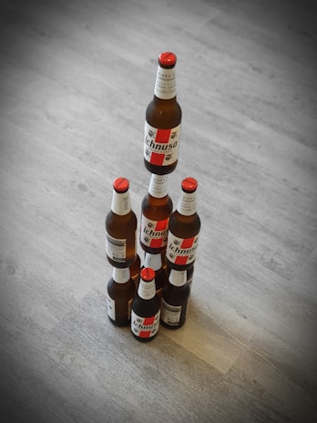 Six beer bottles are stacked in a pyramid formation on a wooden floor. The labels on the bottles have red and black accents, and all the bottles are of the same brand.