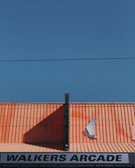 A bright orange corrugated roof with a shadow cast diagonally across it. Attached to the roof is a large, metallic high-heeled shoe decoration. Beneath the roof, there is a sign that reads 'Walkers Arcade'. The clear blue sky serves as a backdrop, with a single horizontal power line running across the image.
