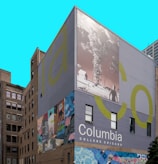 A building facade features large murals, including an abstract art piece with vivid colors and a black-and-white image of a landscape and a tornado. The words 'Columbia College Chicago' are prominently displayed against a solid backdrop.