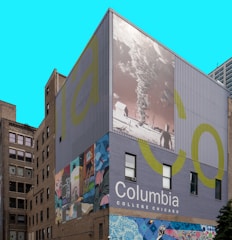 A building facade features large murals, including an abstract art piece with vivid colors and a black-and-white image of a landscape and a tornado. The words 'Columbia College Chicago' are prominently displayed against a solid backdrop.