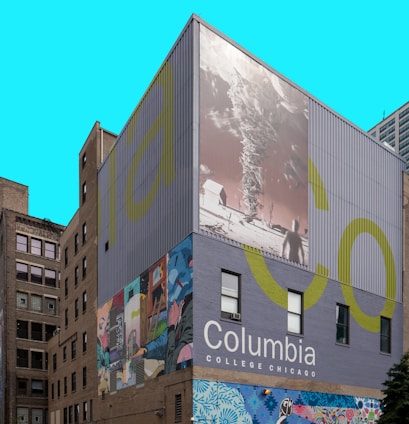 A building facade features large murals, including an abstract art piece with vivid colors and a black-and-white image of a landscape and a tornado. The words 'Columbia College Chicago' are prominently displayed against a solid backdrop.