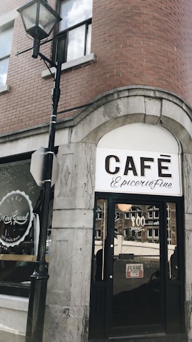 A street-side café with a vintage-style sign reading 'CAFÉ Épicerie Fine' under an archway. The building is made of brick and stone, with a large window displaying 'Chez Mère Grand'. A pole with a lantern is to the left, and there is a 'FERME' sign on the café's glass door.