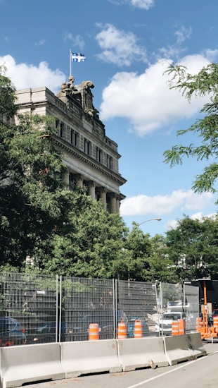 A historical building with classical architectural elements is partially obscured by lush green trees. A flag is visible on top of the building. The foreground features a construction site with safety barriers and orange construction cones. A clear blue sky with a few clouds serves as the backdrop.