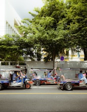 A street scene featuring three tuk-tuks parked along the side of a road. They are occupied by drivers and passengers, with lush green trees and a large building in the background. A no-parking sign is visible nearby.
