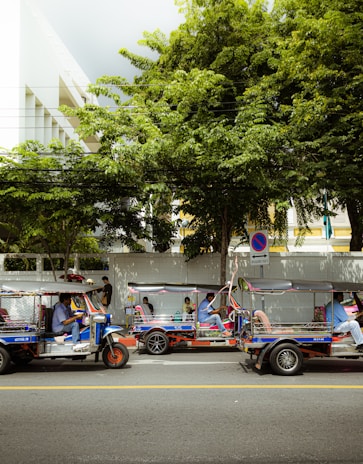 A street scene featuring three tuk-tuks parked along the side of a road. They are occupied by drivers and passengers, with lush green trees and a large building in the background. A no-parking sign is visible nearby.