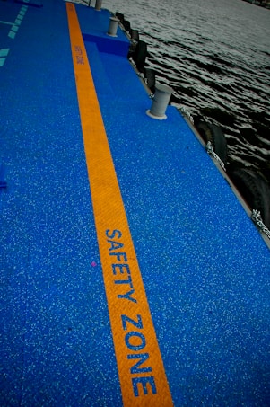 A walkway with a bright blue surface and an orange stripe marked 'SAFETY ZONE' runs alongside a body of water. Several bollards are evenly spaced along the edge, and there are tires attached to the side, likely as bumpers.