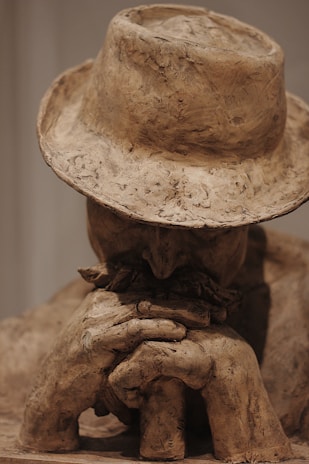 A clay sculpture of a person resting their face on folded hands. The figure wears a wide-brimmed hat, and the overall appearance is rugged and unfinished, with visible texture in the clay.